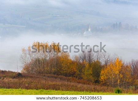 Scenic misty autumn landscape in the Carpathian Mountain with colorful trees on foreground, church in the valley covered by fog. Beautiful nature background.