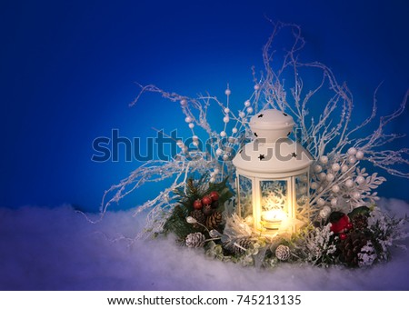 Christmas Eve lantern and decorations background. Picture of a white lantern with lighted candle decorated with pine cones, red berries on a white snow against blue night, evening background. Postcard