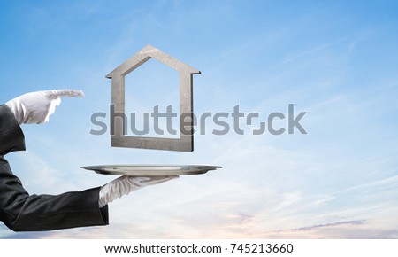 Closeup of waiter's hand in white glove presenting stone house symbol on metal tray and pointing on it with blue cloudy skyscape on background. 3D rendering.