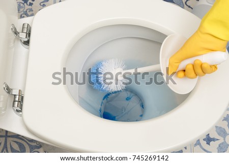 Overhead view of a hand in yellow rubber gloves brushing the interior of a lavatory with blue detergent dissolving in water, janitorial work