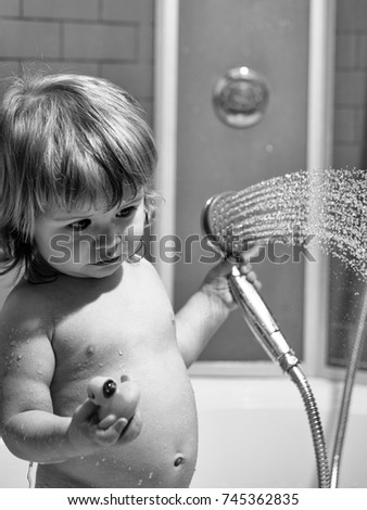 Cute happy smiling funny undressed boy child with blonde curly wet hair taking shower in bath with water indoor playing with toy yellow color duckling, vertical picture