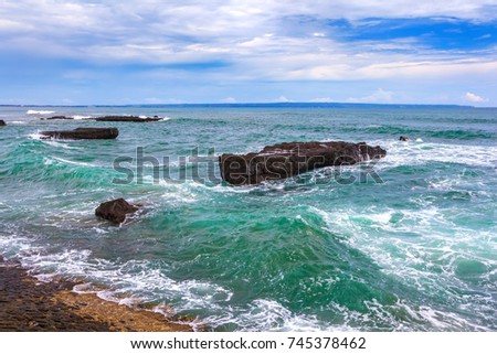 Turquoise sea waves crashing against the rocks roll to the rocky shore forming a foam, a blue sky with white clouds in sunny weather. Beach of Mengwi, North Kuta, Bali, Indonesia.