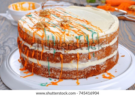 delicious classic carrot cake with cream cheese frosting decorated with walnuts and drizzled with colorful ganache on platter on wooden table with ingredients on background, view from above, close-up