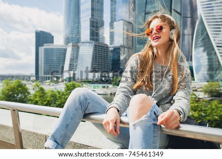 young happy hipster blond woman having fun in city street, happy, urban style, skyscrapers background, freedom, sunglasses, listening to music on headphones, emotional