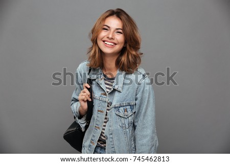 Portrait of a cheery pretty girl in denim jacket holding backpack and looking at camera isolated over gray background