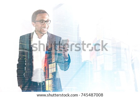 Smiling African American businessman in glasses looking at his smartphone screen and standing in a foggy city. Toned image double exposure mock up