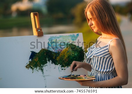 Silhouette of blonde girl paints a painting on the canvas.Palette with paints and spatula, summer sunny day