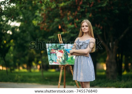 Beautiful girl draws a picture in the park using a palette with paints and a spatula. Easel and canvas with a picture.