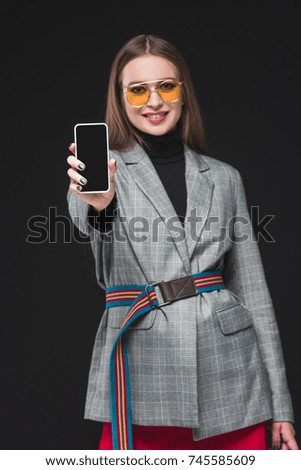 Smiling woman in autumn outfit showing smartphone isolated on black