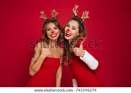 Picture of happy young women friends wearing christmas deer costumes standing isolated over burgundy background wall. Looking at camera showing thumbs up.
