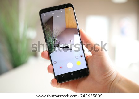 A hand holding a modern generic mobile phone which displays the camera app on the touch screen. The image is taken in the living room of a bright home, full of light and plants.