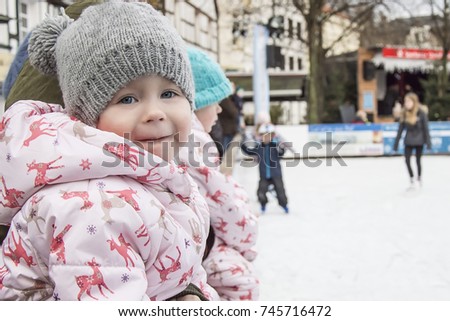 Little cute little girl in gray cap watches children on the ice in winter.
