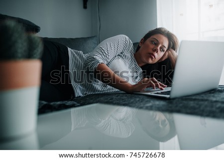 A young woman is using her laptop while lying on her couch.