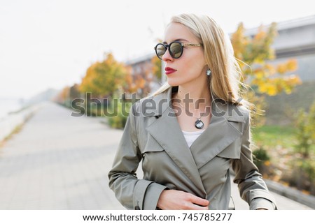 Close-up portrait of stylish pretty blonde with long hair dressed in autumn coat and black sunglasses with vine lipstick is posing at the camera outside. Romantic girl on the street with pretty smile