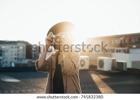 Trendy and attractive beautiful woman with analog vintage film camera makes photos on rooftop at sunset time, creates content for social media channels and applications, popular influencer