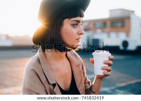 Natural beauty pretty woman with short haircut and in black fedora hipster hat, sips on coffee or tea from blank white mockup take away to go cup, on warm summer evening spring sunset in big city Royalty-Free Stock Photo #745832419