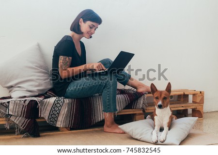 Pretty woman works from home or startup coworking space, sits barefoot on bench and writes code or blog on laptop, her best friend dog puppy lays next to her on pillow Royalty-Free Stock Photo #745832545