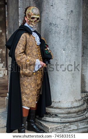 Gold colonial Venice carnival costume and skull mask with green cape and marble column in background