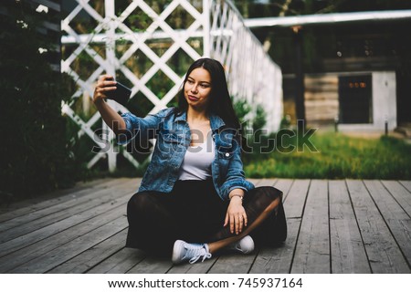 Stylish young woman talking on video chat via modern smartphone device sitting outdoors.Charming with brunette hair female making selfie photos on modern telephone for publication on internet website