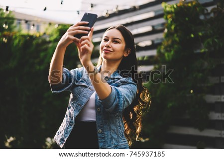 Attractive female with brunette hair looking at front camera and making selfies on modern smartphone device.Smiling young woman in stylish denim jacket taking photos on telephone standing outdoors