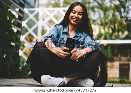 Cheerful young woman laughing and talking with friend in video chat via smartphone device sitting in the lotus position.Positive hipster girl making photos on mobile phone to publicating new post