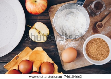 Ingredients for apple pie cooking with fresh red apples, flour, eggs, brown sugar and spices on a rustic wooden background.