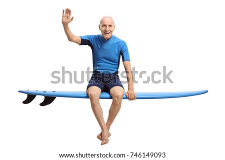 Elderly man in a wetsuit sitting on a surfboard and waving at the camera isolated on white background