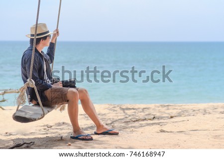 Travel lifestyle in summer season. Young Asian man photographer sitting on wooden swing at tropical beach. Male traveler enjoy looking at seascape scenery of Thailand. Relaxation and carefree concepts