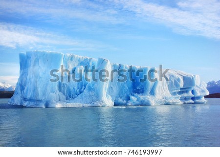 Single big blue iceberg in blue water in the center of the picture and blue sky with clouds on the background