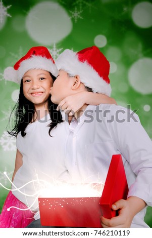 Happy little boy holding a Christmas gift and kissing his sister while wearing Santa hat in front of bokeh lights background