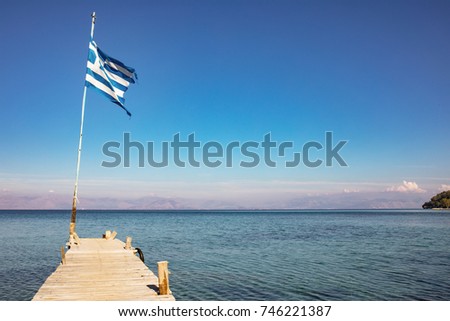 Greek flag on the end of a wooden jetty or a pier on the Ionian Sea - a part of Mediterranean Sea, dark blue sky, autumn in Greece, Ionian Islands, Corfu. Albania in background.