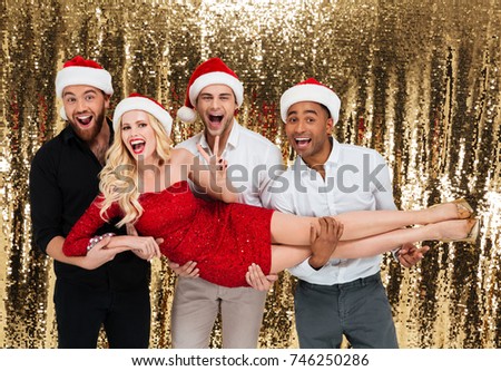 Image of happy group of friends wearing christmas hats standing over glitter bright background with their woman friend on hands. Looking camera.