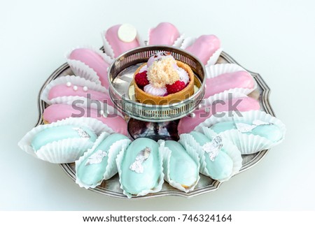 Elegant french pastry glazed pink and mint eclairs decorated with white edible balls on a silver plate on a white background with a fruit tartal. Party sweets.