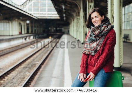 Beautiful woman wears knitted sweater and scarf, sits on suitcase near platform, holds smart phone in hands, waits for train, going to travel in other country, Female traveler with happy expression