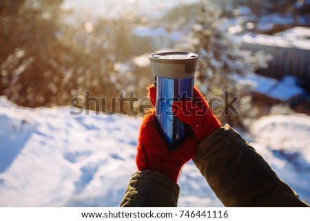 Thermos with drinks in woman's hand with red mittens. Blue thermos cup with a straw. Sunny day.