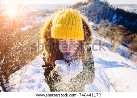 happiness, winter holidays, tourism, travel and people concept - smiling young woman in yellow knitting hat and mittens blowing snow on the hill over snowy city view background.
