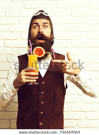 handsome bearded aviator man with long beard and mustache on surprised face holding glass of alcoholic cocktail in vintage suede leather waistcoat with hat and glasses on white brick wall background