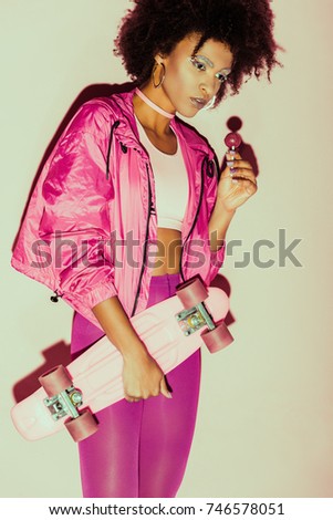 african american 80s girl in pink clothes posing with penny board and lollipop for fashion shoot