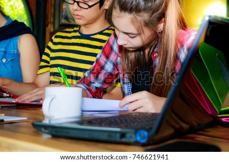 Education concept: A beautiful student girl is reading and writing something on the paper. Charming girl is doing her homework or report with her friends at some cafeteria with notebook computer