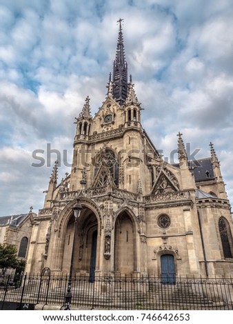 Beautiful dramatic church photograph of Saint Bernard Chapel with intricate Gothic architectural detailing and dark colored spire in Paris France with fluffy white clouds in a blue sky above