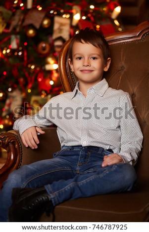 Smiling boy in jeans and shirt sitting near Christmas tree. Family Holidays