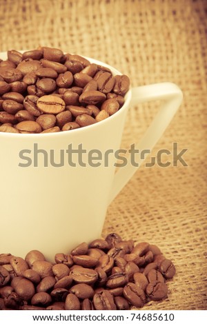 This is a shot of roasted coffee beans inside and at the base of a coffee cup with a burlap sack background. Shot with a shallow depth of field in a warm retro tone with vignetting.