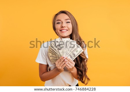 Portrait of a happy satisfied girl holding bunch of money banknotes and looking at camera isolated over yellow background