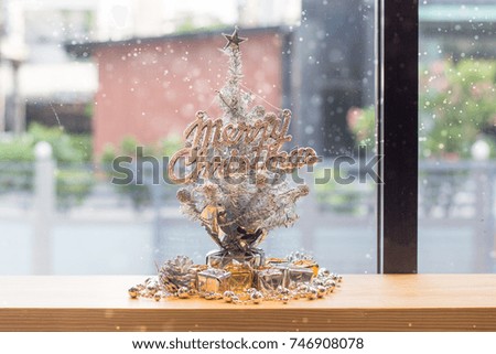 A silver Christmas tree is on the wooden table decorated with gift boxes, pines and snowflake. "Merry Christmas" text is decorated on the Christmas tree.