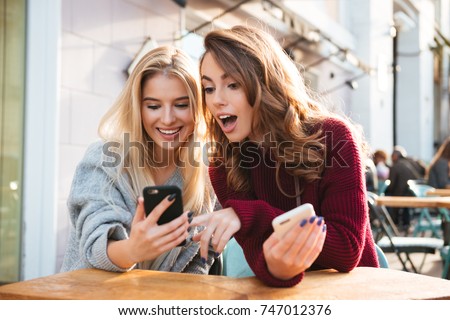 Two excited young girls using mobile phones while sitting at the cafe outdoors and pointing finger Royalty-Free Stock Photo #747012376