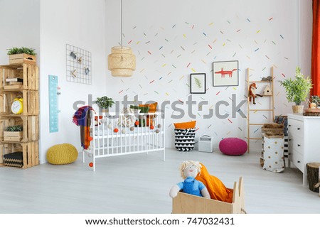 Doll in wooden box in spacious scandi baby's room interior with pink and yellow pouf and white bed