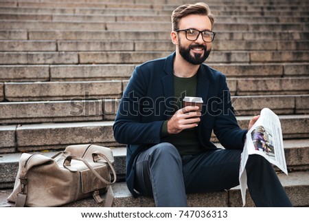 Picture of happy young bearded man sitting outdoors on steps reading newspaper and drinking coffee. Looking aside.