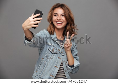 Funny teen girl in jeans jacket showing peace gesture while taking selfie on smartphone, isolated on gray background