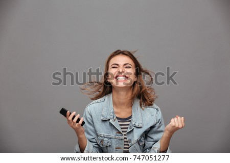 Close-up portrait of beautiful screaming young girl holding smartphone showing winner gesture, isolated on gray background Royalty-Free Stock Photo #747051277