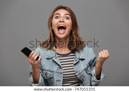 Overjoyed girl in jeans jacket holding mobile phone and showing winner emotions, looking at camera, isolated on gray background